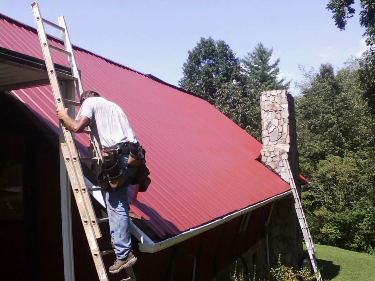 Roofer on a ladder beside a red metal roof and stone chimney — Soffit & Fascia Repair by Roofing Brothers in Schererville